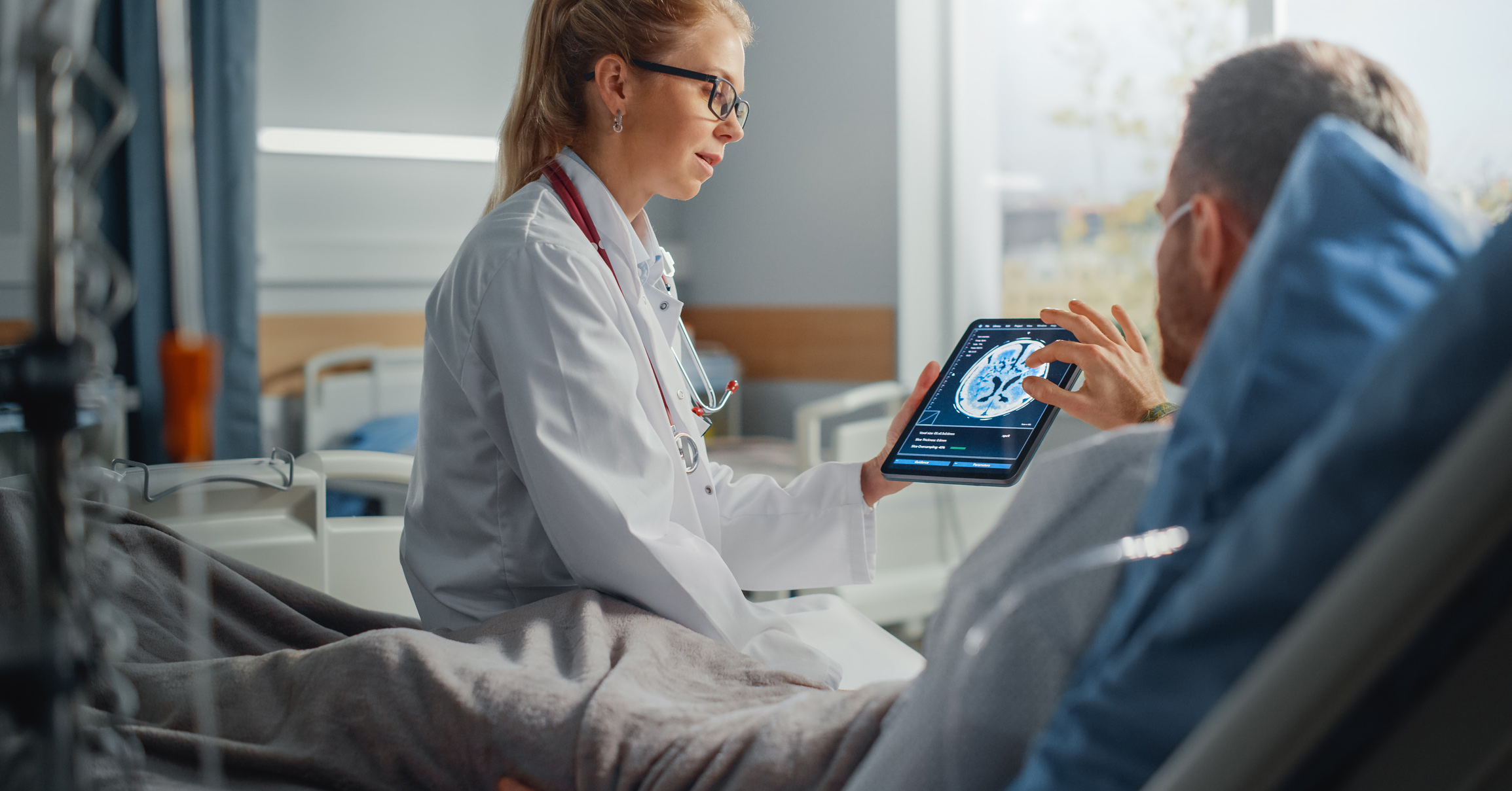 A female doctor sat on a patients bed showing him a AI generated brain scan on an ipad