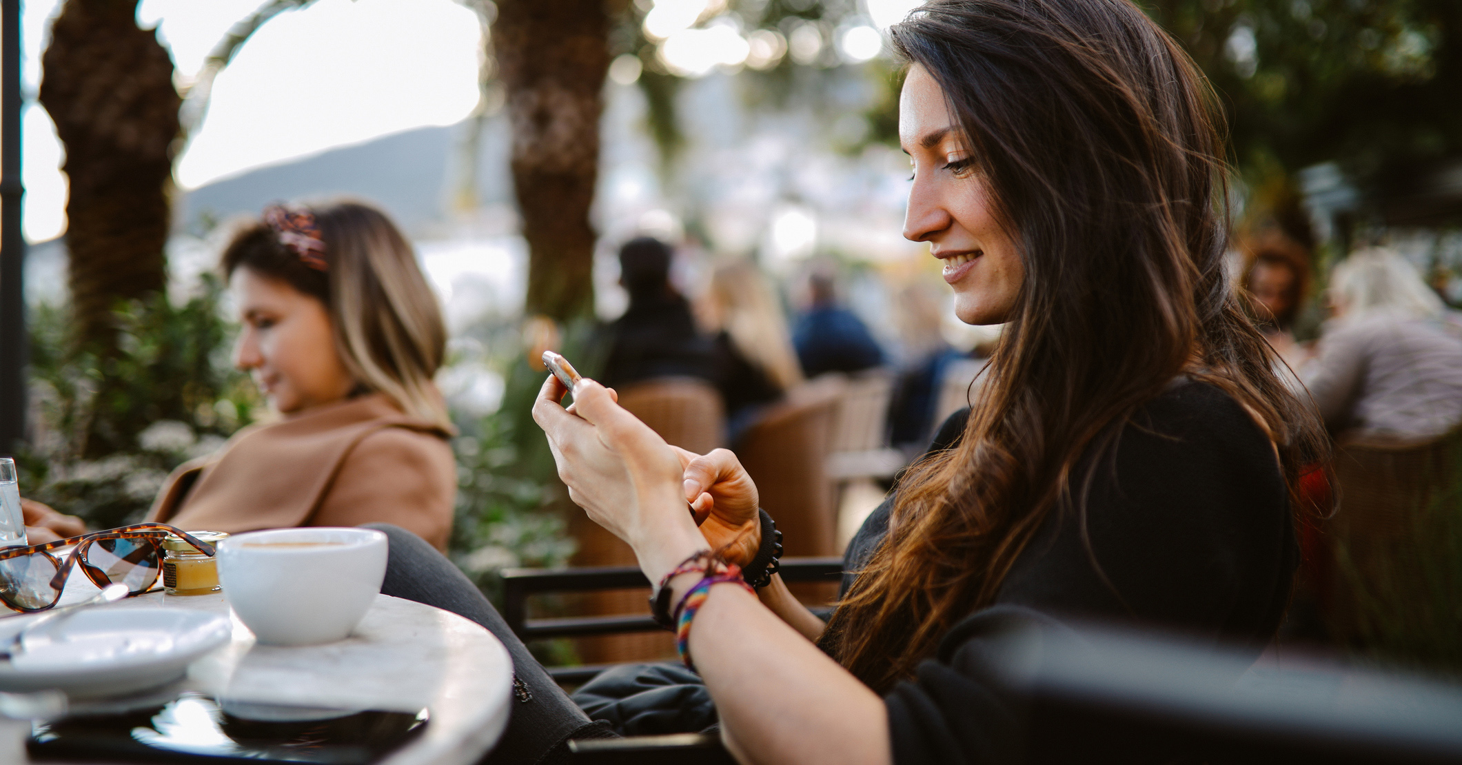 A woman sat outdoors in a busy cafe smiling at her phone with a coffee