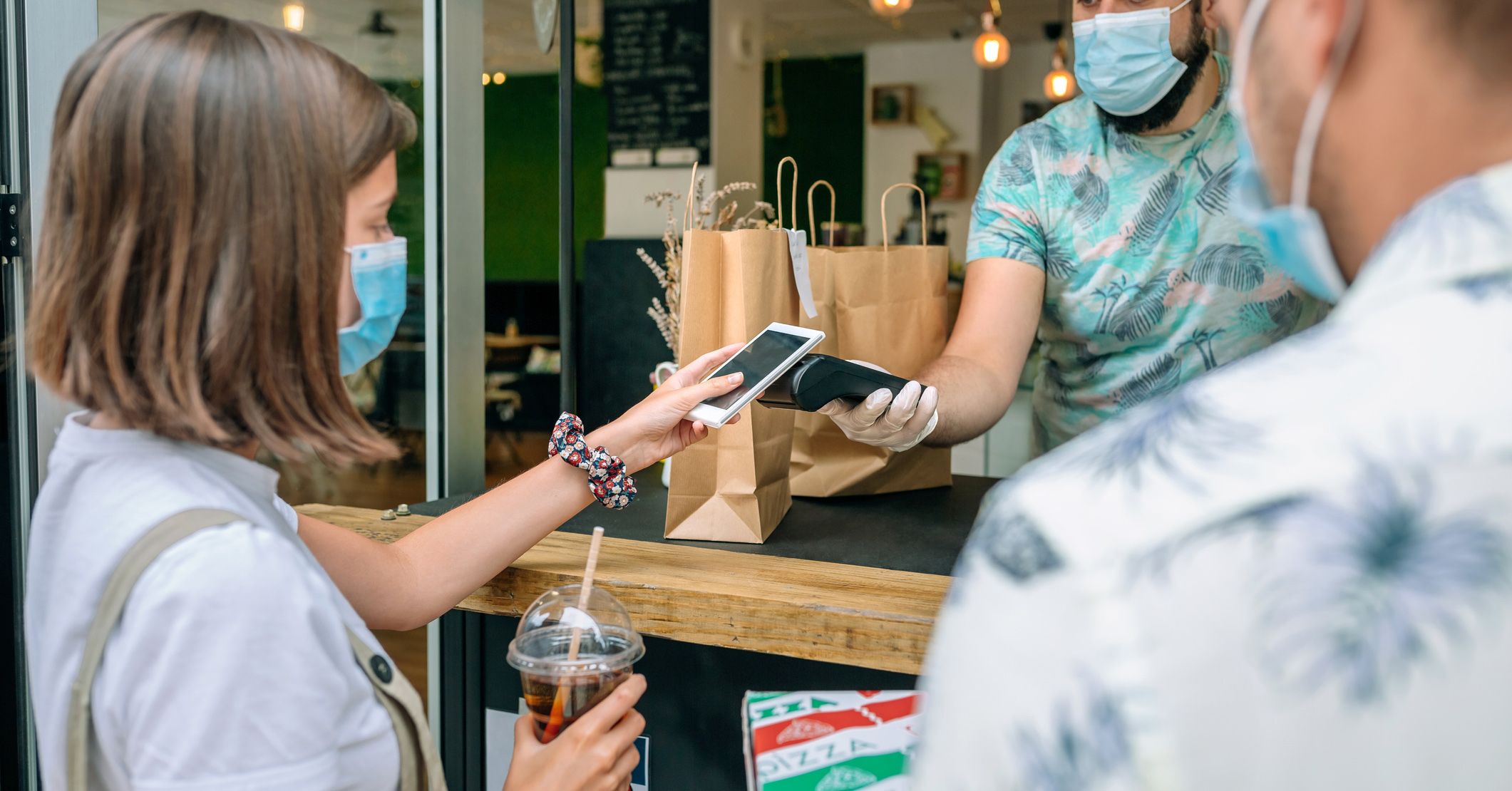 A woman wearing a mask holding a coffee paying for shopping using her phone